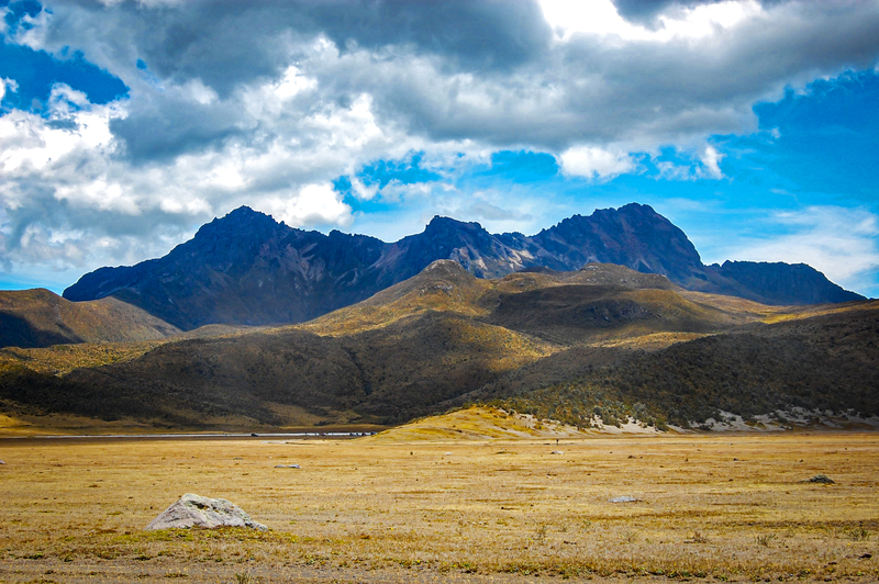 Image of low, craggy volcano peak across brown field under cloudy blue sky