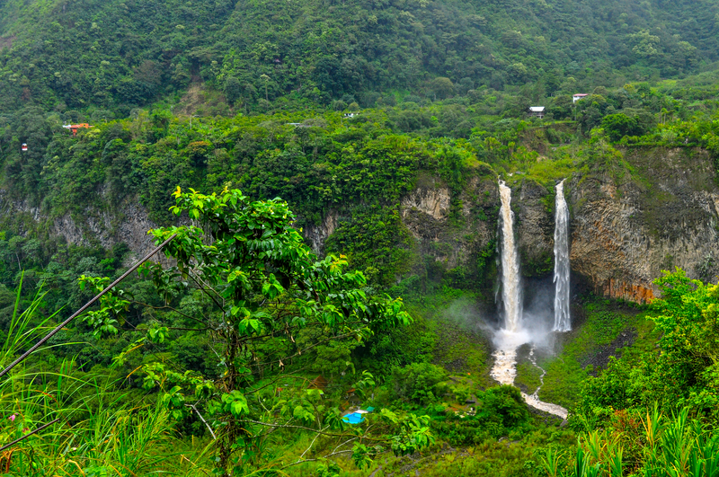 Far-away shot of two tall, thin waterfalls cascading down a cliff in lush rainforest.