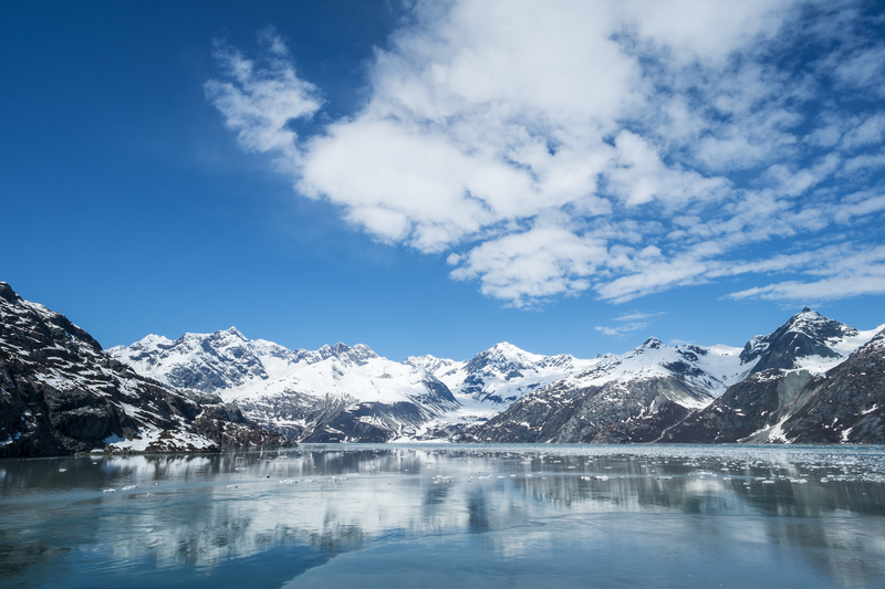 Image of snowcapped mountains and glacier under blue sky reflected in calm bay dotted with icebergs.