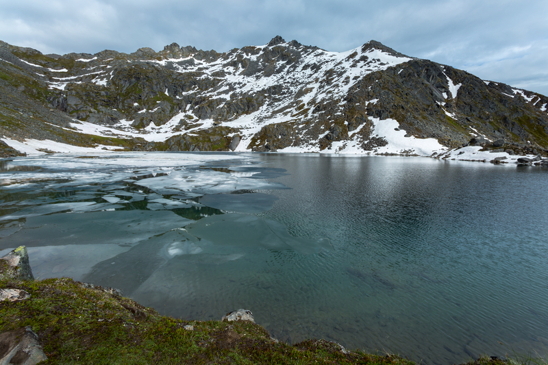 Partially frozen alpine lake ringed with low snowy mountain.