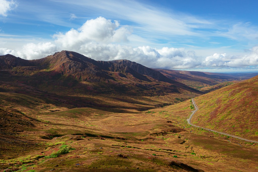 Mountainous, reddish-brown landscape with winding road under blue cloudy sky.