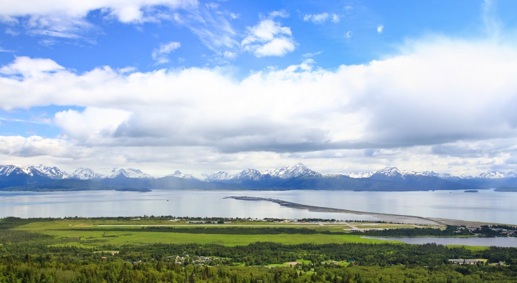 Landscape view of Homer Alaska with green land stretching to calm bay with snowcapped mountains in the background