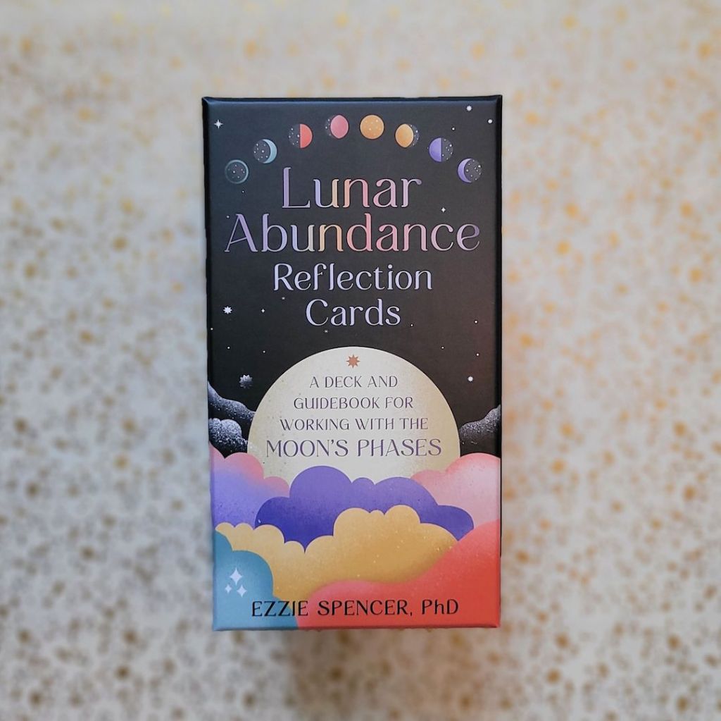 Photo of the “Lunar Abundance Reflection Cards: A Deck and Guidebook for Working with the Moon’s Phases” box laid above a gold and white backdrop