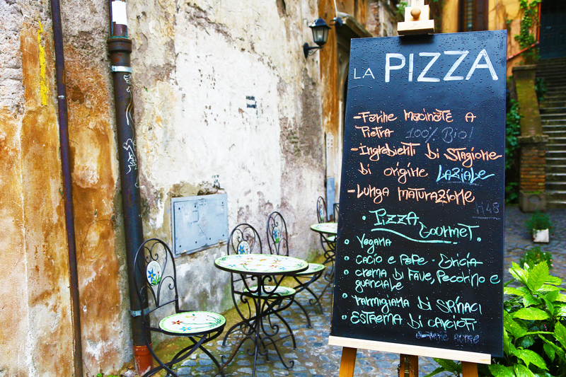 Small metal cafe tables at an outdoor restaurant with a blackboard menu listing kinds of pizza