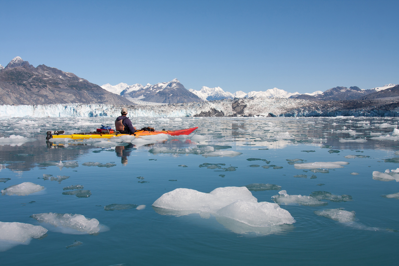 A kayaker in a yellow and orange kayak in water dotted with small icebergs with a glacier in the distance.