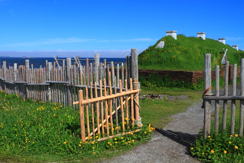 A wooden fence with an open gate and a path leading to a lush green hill with chimneys sticking out of it