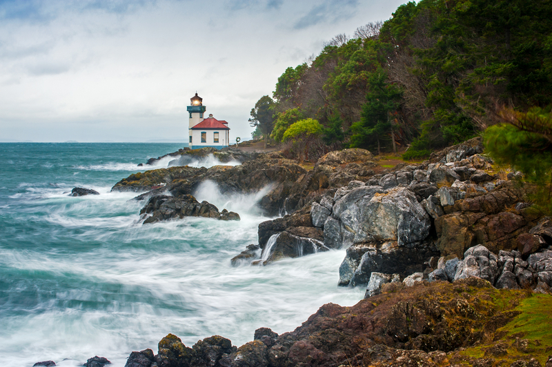 Image of a red and white lighthouse on a rocky promontory with crashing turquoise waves.