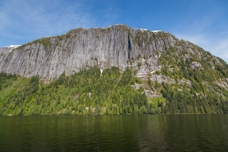 Image of tall stone cliffs with trees at the base rising up from calm dark green water under bright blue sky