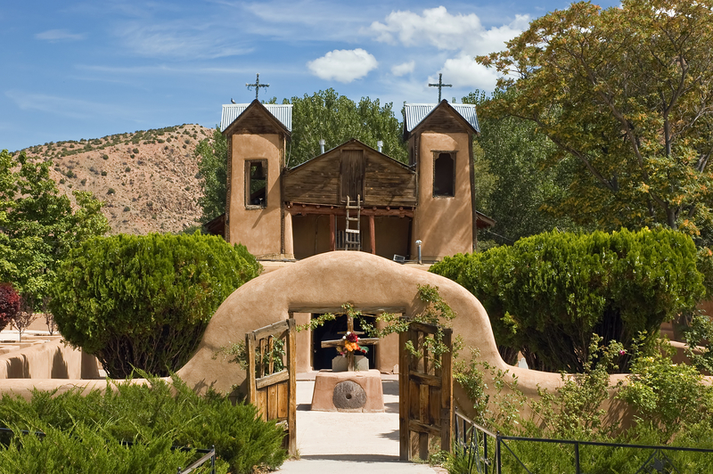 Image of archway leading to adobe church surrounded by gardens.