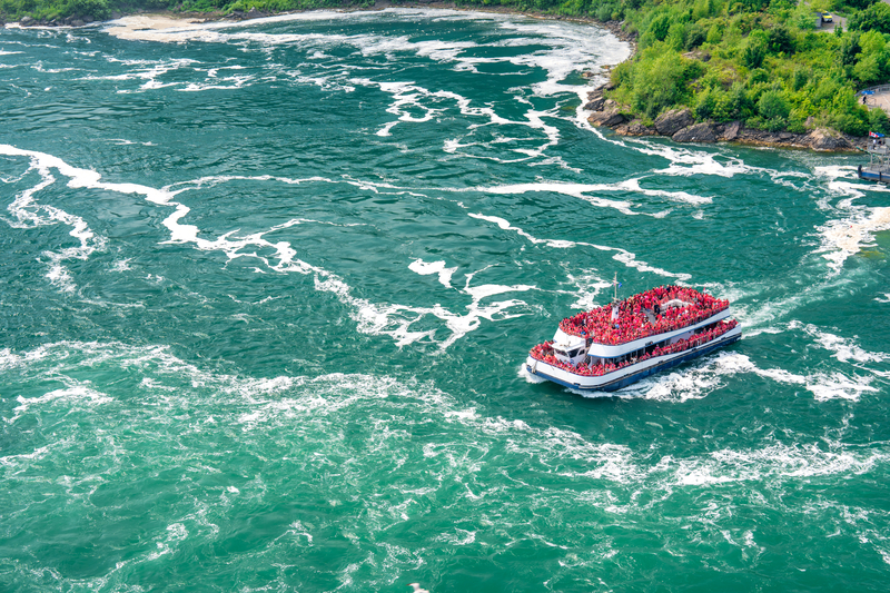 Aerial view of red, two story boat with passengers on foamy, turquoise blue water.