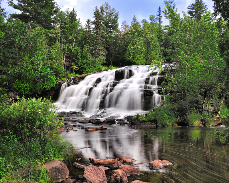 Image of cascading waterfalls into a pond surrounded by tall green trees.