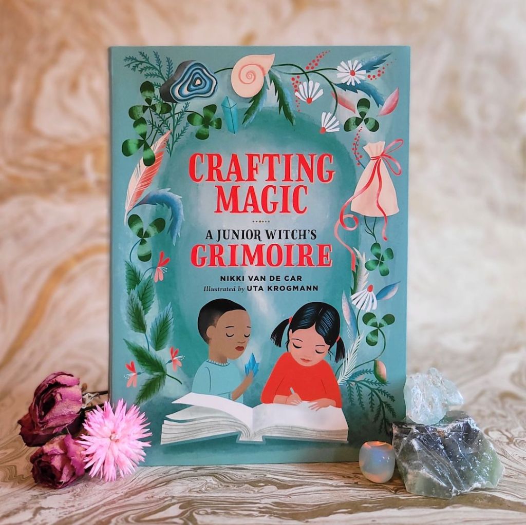 Photo of “Crafting Magic: A Junior Witch’s Grimoire” standing next to flowers and crystals in front of a gold marble background