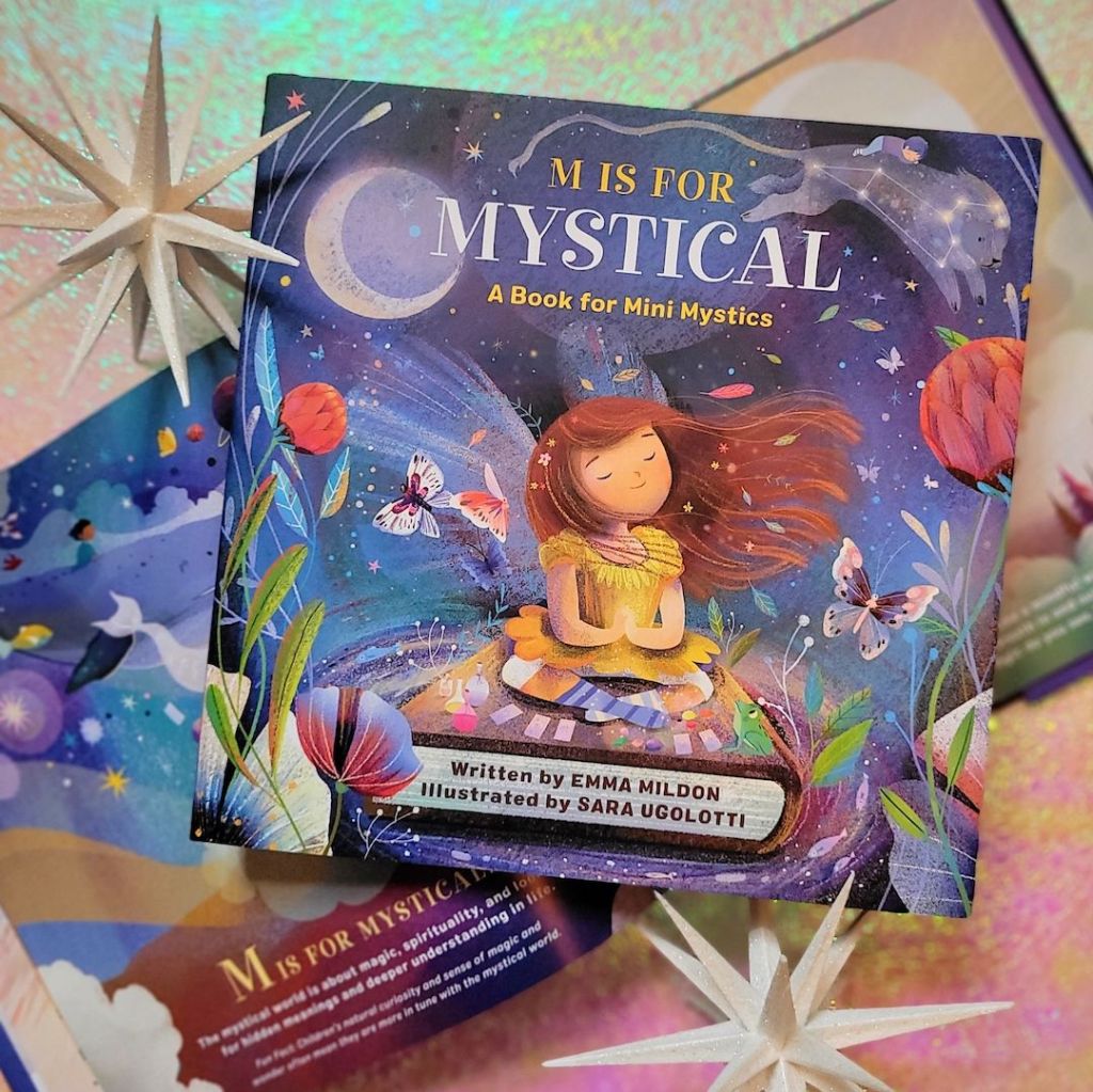 Photo of a closed copy of “M is For Mystical: A Book for Mini Mystics” laid above an open copy of the book. The stack sits between decorative white starbursts on a pink and green iridescent backdrop.