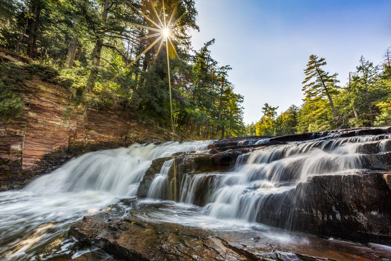 Image of silky waterfalls rushing over rocks with sunbeams shining through trees lining the river.