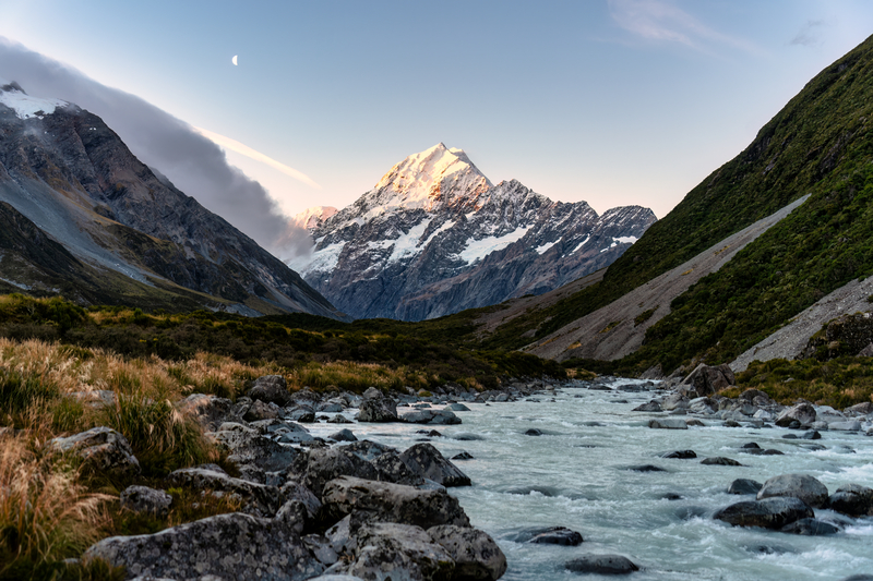 Image of river rushing over rocks in an alpine valley with a glowing, snow-capped mountain in the distance.