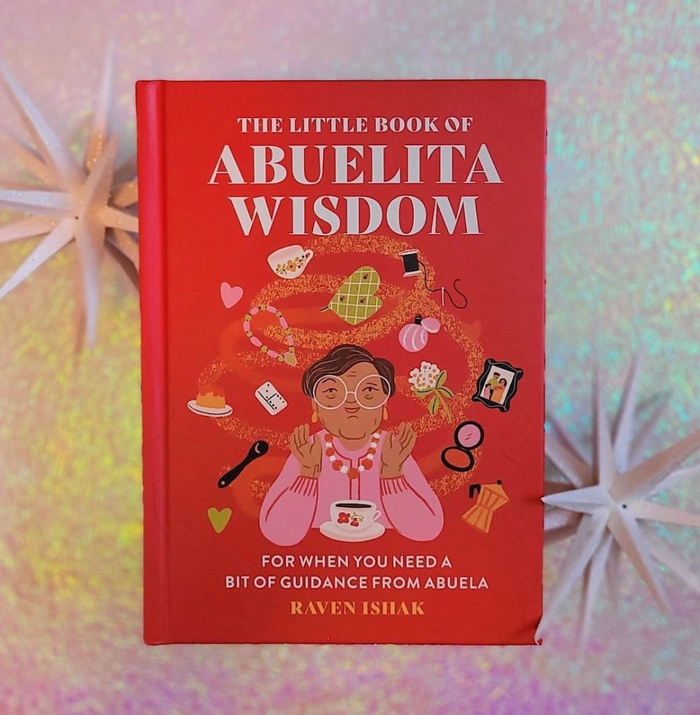 Photo of “The Little Book of Abuelita Wisdom” laid next to two white decorative starbursts against a pink and green iridescent background