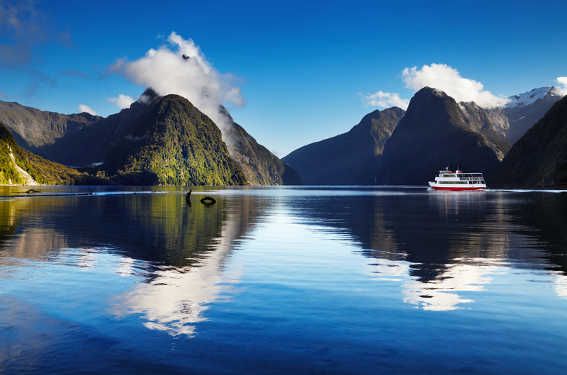 Image of green mountains capped with white whispy clouds reflected in calm blue water.