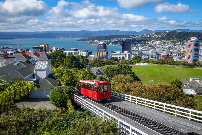 Image of red cable car climbing up a grassy green hill with city skyline and ocean in the distance.