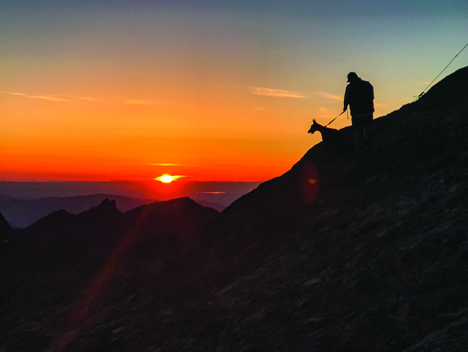 A hiker and his dog on the side of a mountain silhouetted against glowing orange sunset.