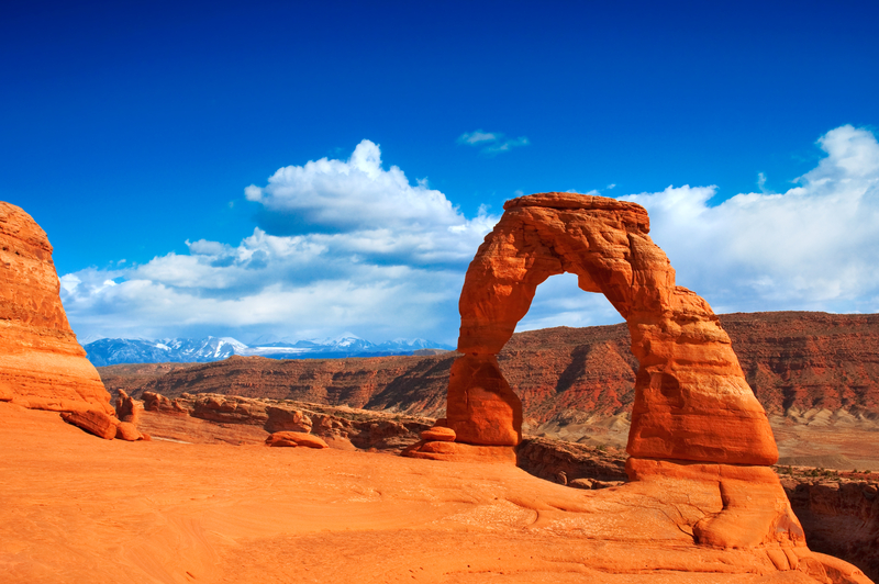 Image of red-orange rock formation in the shape of an arch with red mountains in the background under bright blue sky.