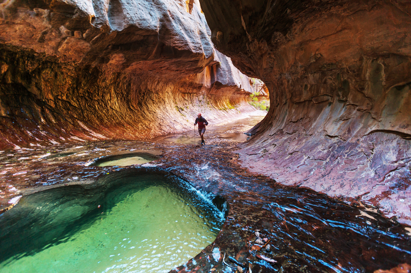 A hiker treks through a shallow turquoise river in a red rock canyon.