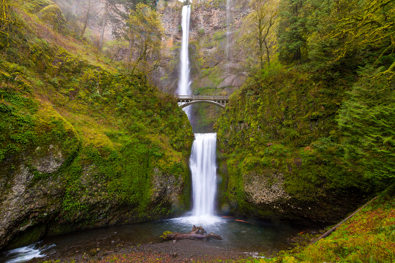 Two waterfalls cascade down a tall, mossy green gorge with a bridge stretching across the lower falls.