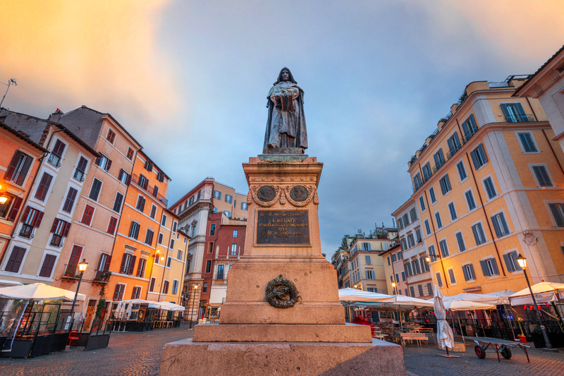 Image of a tall statue with a metal figure on it in the center of a plaza lined with pastel-hued buildings softly illuminated at dawn 