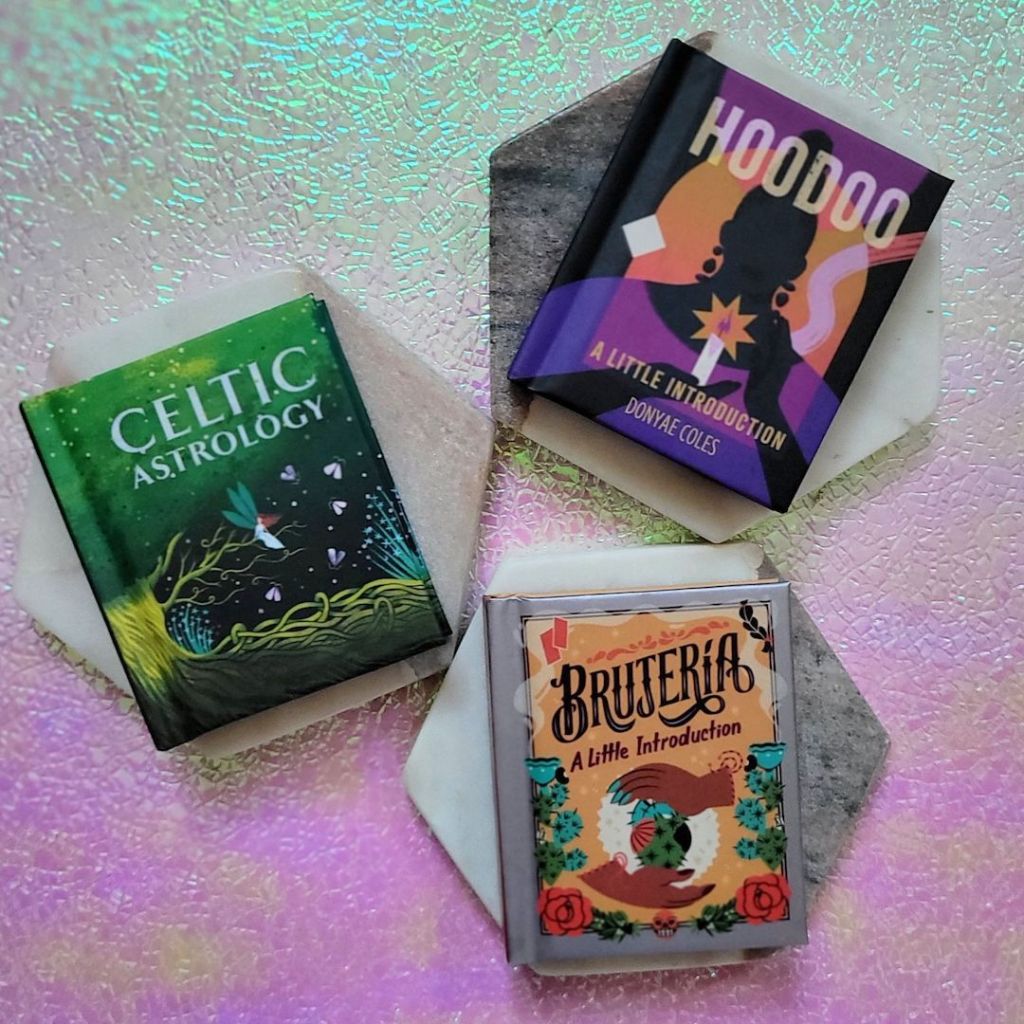 Photo of “Brujería: A Little Introduction,” “Hoodoo: A Little Introduction,” and “Celtic Astrology” laid on marble hexagons above a pink and green iridescent background