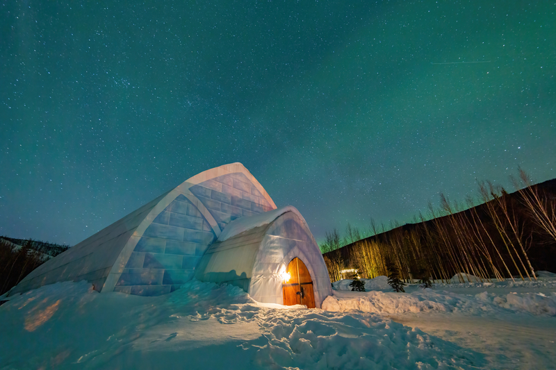 Green and blue northern lights over an igloo-like structure and snowy landscape.