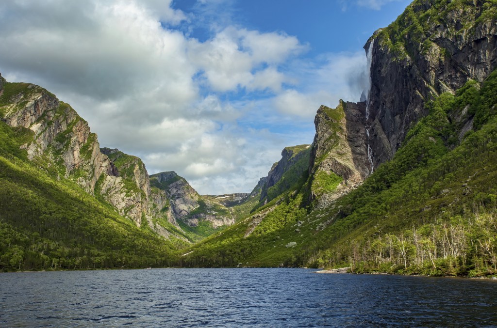 Image of towering, green rocky mountains overlooking bay of calm water.