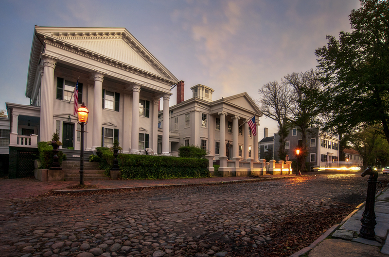 White houses with tall classical columns along a cobblestone street at dusk with illuminated street lamps.